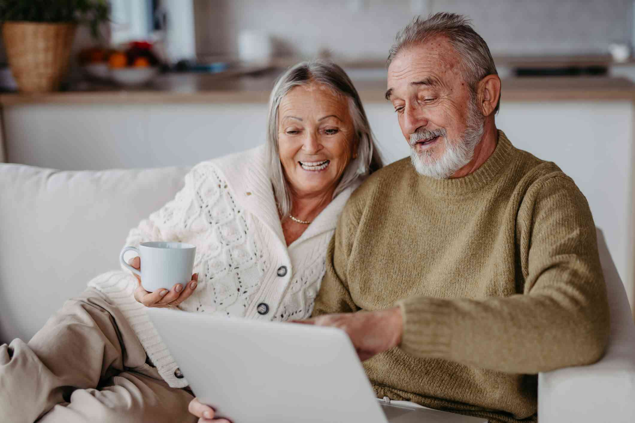 9f64103c026470e8f7bda0293685d79b-senior-couple-look-at-a-laptop-while-smiling-on-the-couch-together_l-1