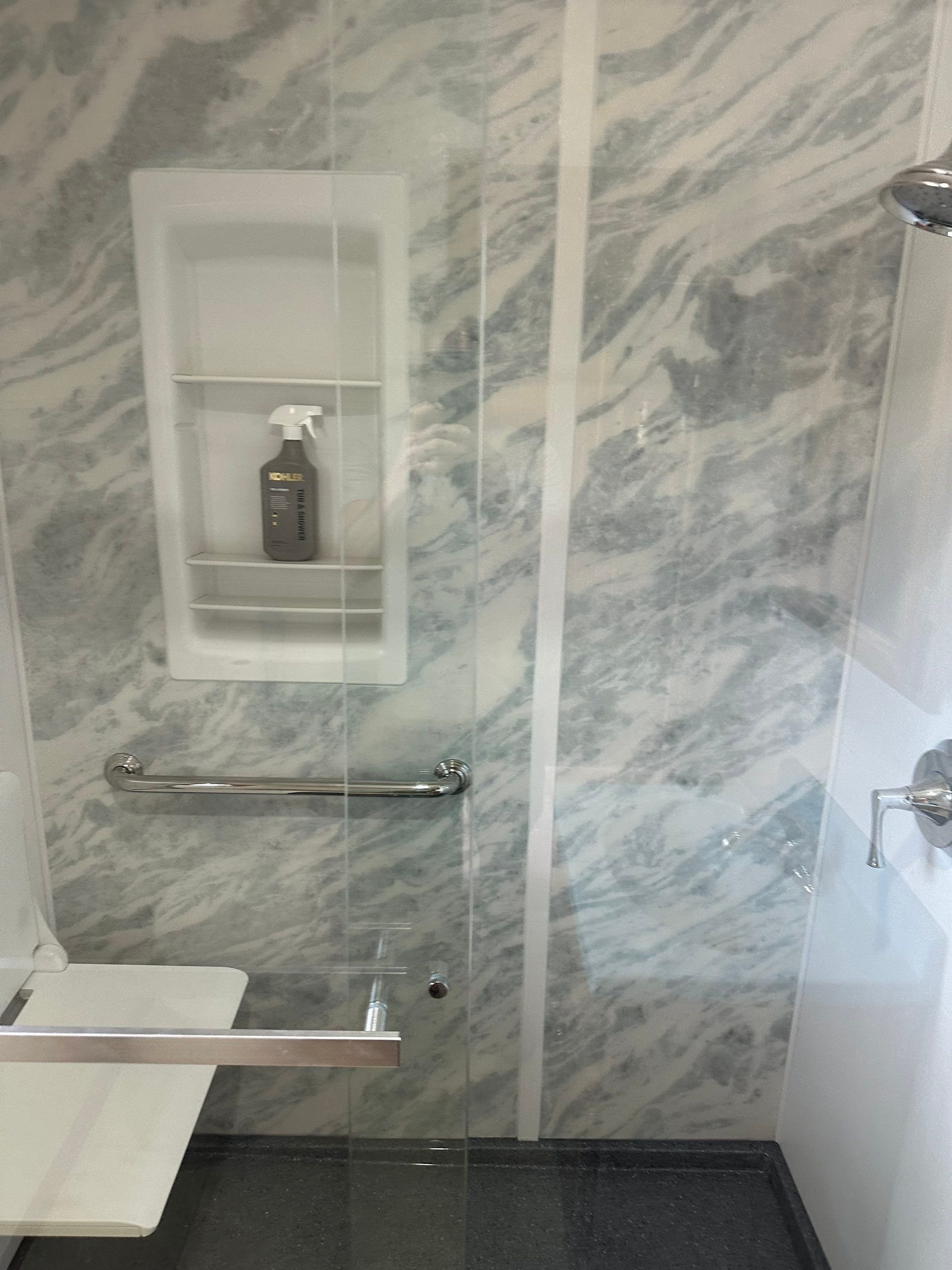 After photo of a Kohler walk-in shower remodel with marble-style LuxStone walls, built-in shelving, grab bar, and glass door for safe, modern bathroom design.