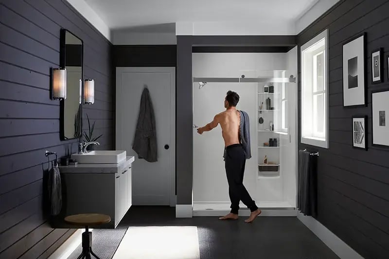 Man stepping into a KOHLER walk-in shower in a dark modern farmhouse bathroom with shiplap walls and matte black fixtures.