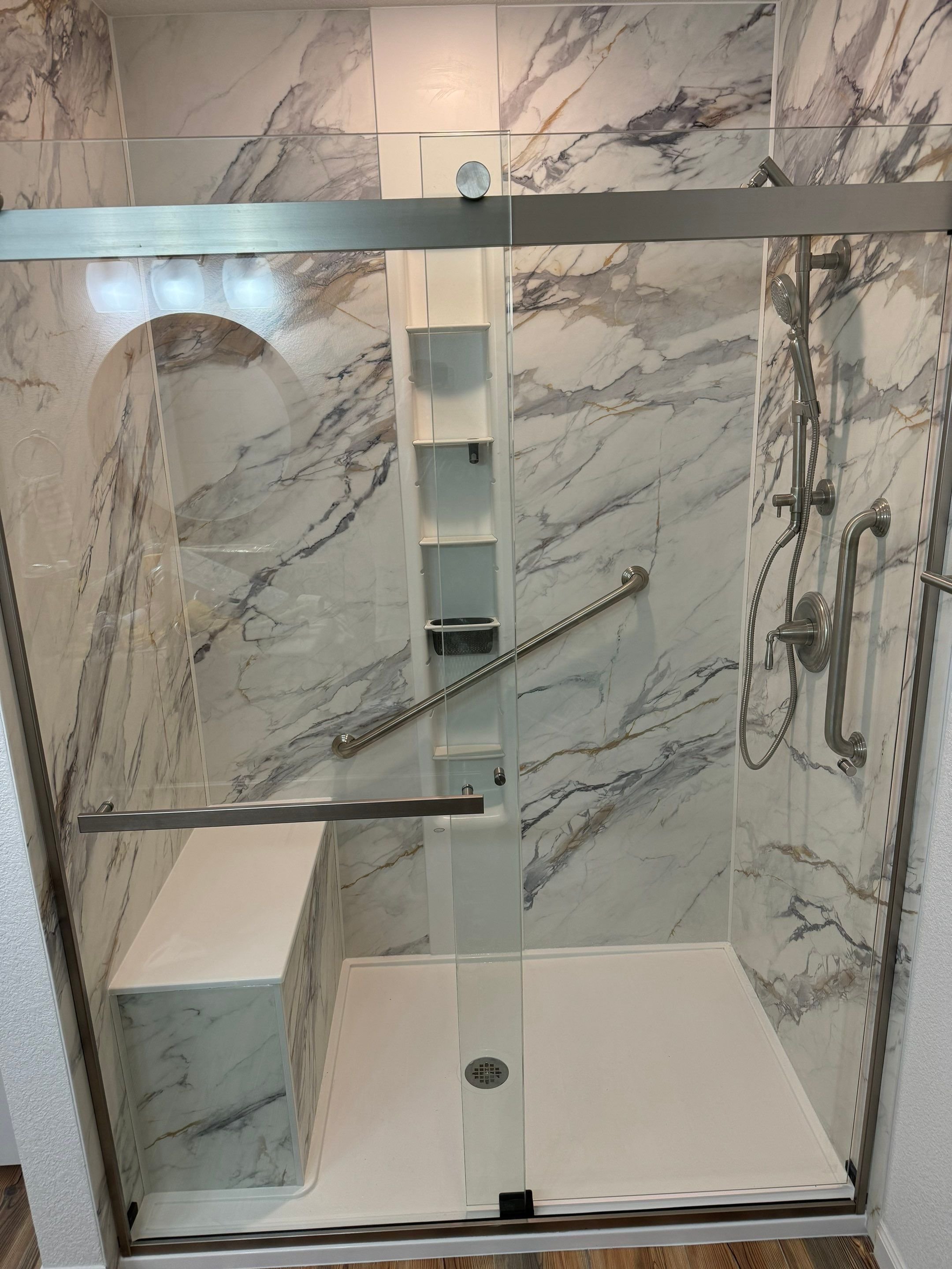 Cheyenne bathroom remodel on Bradney Avenue with marble-look wall panels, built-in shelving, grab bar, and frameless glass shower door