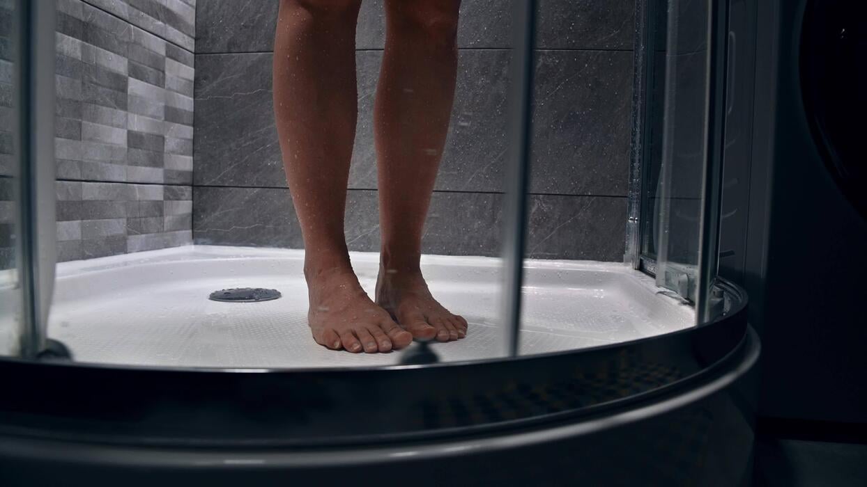 Bare feet of a person standing in a modern shower enclosure with glass doors, water droplets visible on the floor and tiled walls creating a clean, refreshing atmosphere