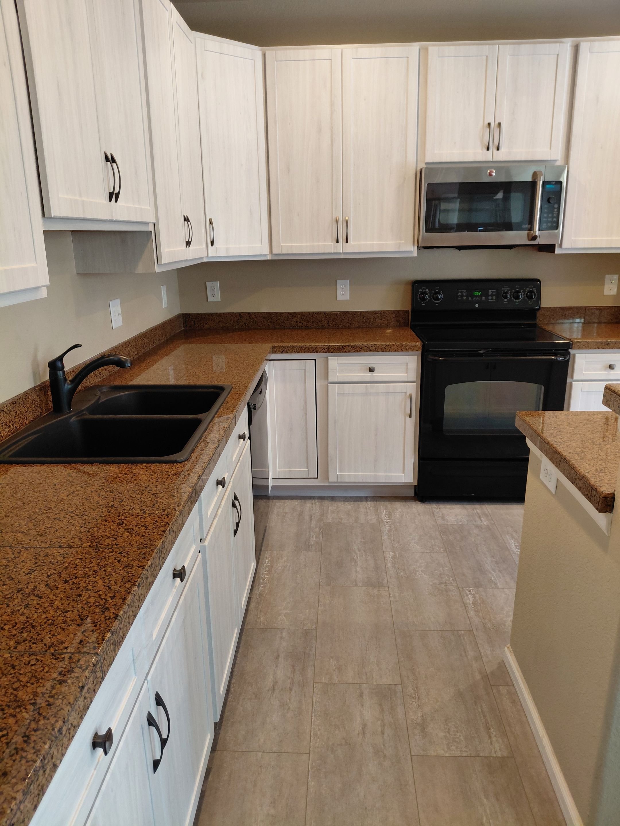 Light woodgrain kitchen cabinet restyling in Alabaster, CO with brown granite countertops and black appliances