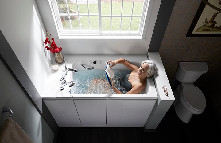 Senior woman relaxing and reading in a modern white walk-in bathtub with water jets, under a large window in a bright, accessible bathroom.