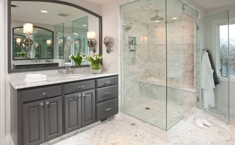 Modern bathroom remodel with frameless glass walk-in shower, marble tile walls, dark gray vanity, and large mirror with elegant lighting fixtures.
