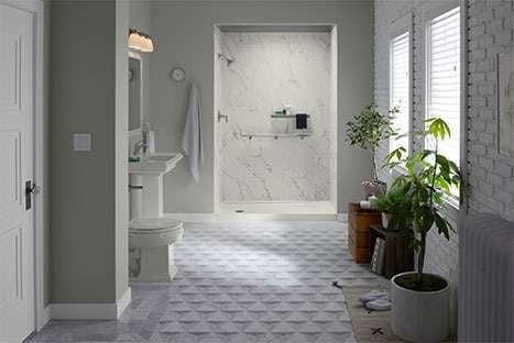 Modern bathroom with open walk-in shower featuring marble-look walls, pedestal sink, geometric tile flooring, and natural light with potted plants.