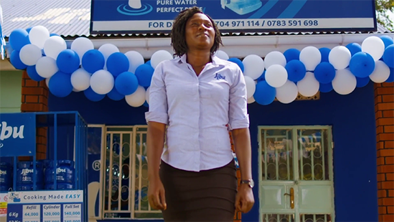 Mrs. Musoke, a Jibu franchise owner in Africa, stands proudly outside her clean water retail storefront decorated with blue and white balloons, symbolizing community impact and access to safe drinking water.