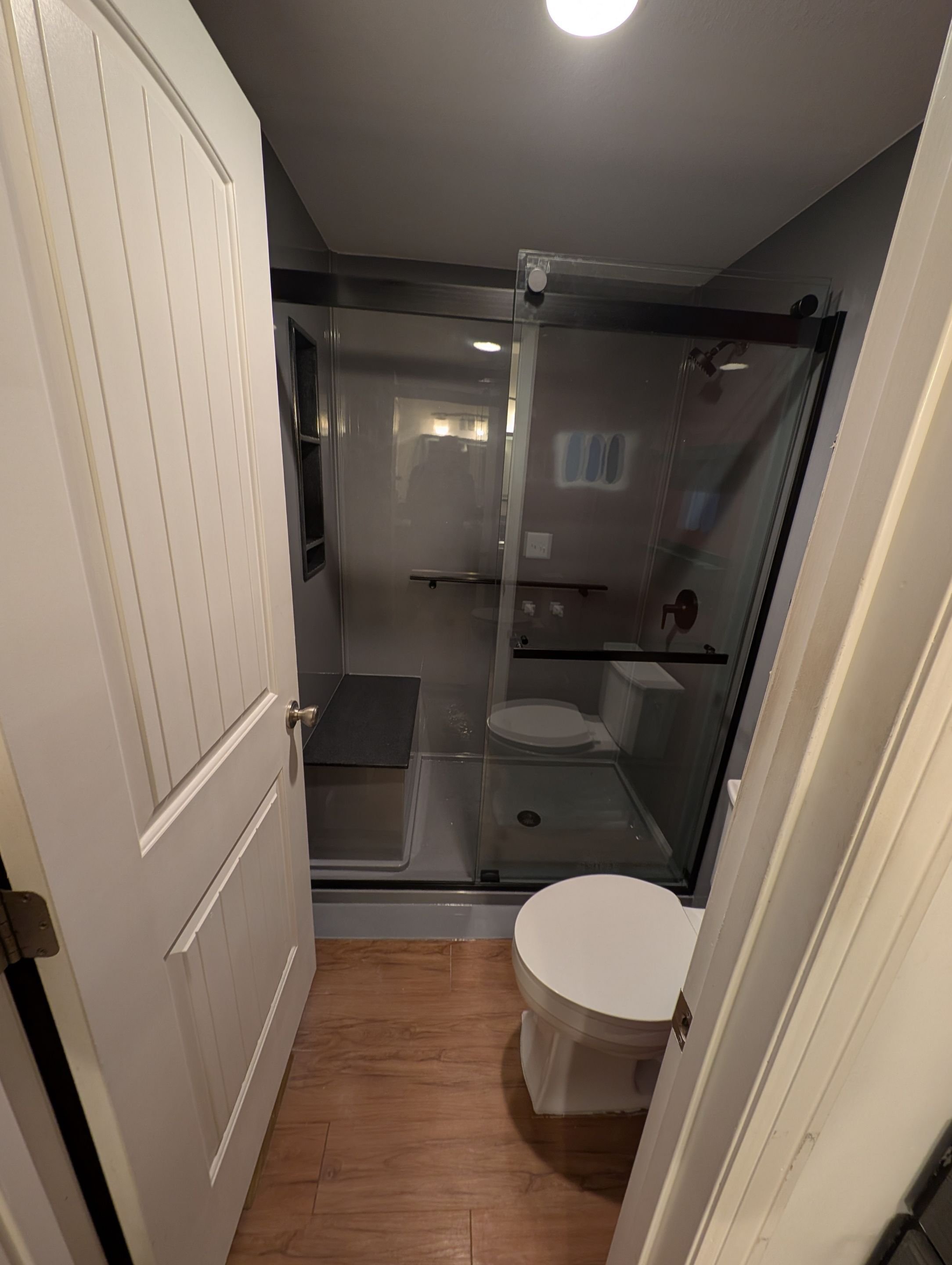 Compact walk-in shower renovation in Aurora, CO featuring sliding glass doors, built-in bench seating, and wood-look flooring